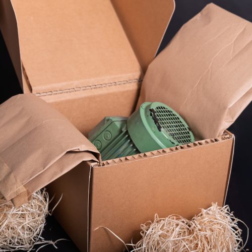 Fibre de bois naturelle en vrac et en enveloppe papier utilisée comme calage écologique dans des cartons, protégeant une pièce industrielle pendant le transport.